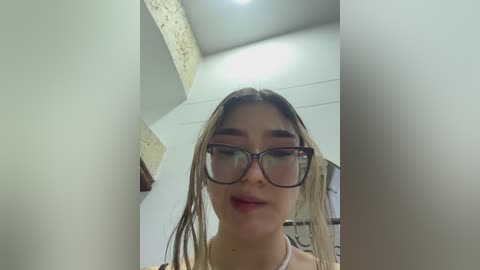 Media: Video of a young woman with long blonde hair, wearing large black-framed glasses, a light-colored top, and a necklace. She stands in a beige-walled room with textured ceiling and a bright light above.