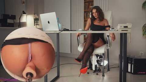 Video of a curvaceous woman with dark hair, wearing black lingerie, sitting at a desk in a modern office. Close-up inset of her buttocks, showing a large dildo.