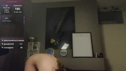 A dimly-lit video captures a partially nude person with fair skin, bending over a table. The room features a large framed poster and a whiteboard on beige walls.