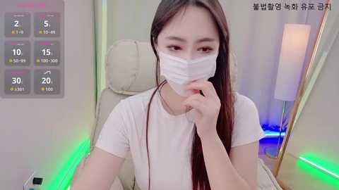 A video of an Asian woman with long dark hair, wearing a white mask, sitting in a beige chair with a modern lamp and green LED lights in the background.