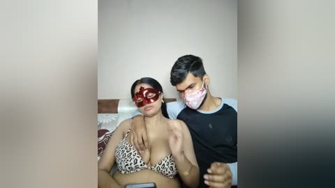 Video of a South Asian woman in a leopard print bikini, wearing a red mask, and a man in a black shirt, both indoors, with a blurred background.