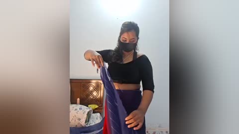 Video of a woman with medium-dark skin, wearing a black mask, black long-sleeve top, and blue skirt, sorting laundry in a small room with a wooden cabinet and a patterned pillow.
