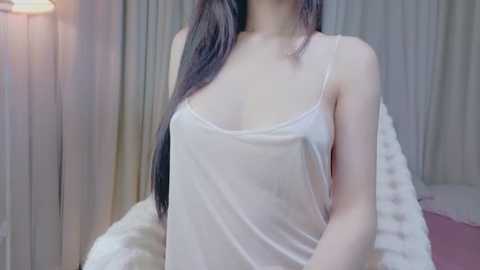 A video of an East Asian woman with long, straight black hair, wearing a sheer white tank top, posing against a backdrop of beige curtains.