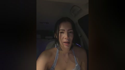 A dimly lit video shows a young woman with medium brown skin, dark hair, and a light blue halter top, seated in a car with a slightly blurred background.
