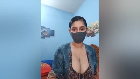 Video of a light-skinned woman with dark hair, wearing a black face mask and a low-cut, patterned dress with a blue cardigan. She's in a light blue room with floral art on the wall, and a bed with red and orange blankets.