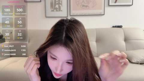 Video of an East Asian woman with long, straight brown hair, wearing a black top, sitting on a beige couch. She has a neutral expression. The background features framed artwork and a digital screen displaying social media statistics.