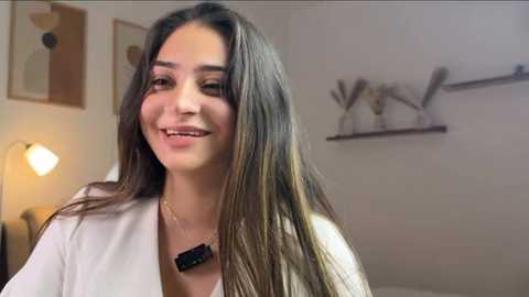 Video of a smiling woman with long, straight brown hair, wearing a white robe, standing in a modern, minimalist room with white walls, abstract art, and a shelf with decorative items.