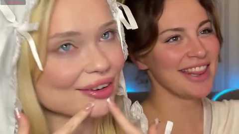 Video of two young women with light skin, one blonde and one brunette, smiling, applying makeup. They wear white headbands with lace details.