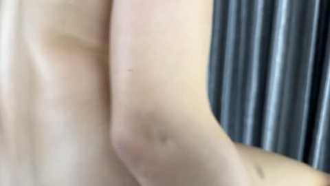 Video of a nude, light-skinned person's back and arm against a dark, vertical curtain background. The skin is smooth, with slight shadows indicating natural light source. The image is close-up, focusing on the texture and contours of the body.