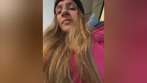 Video of a young woman with long, wavy, light brown hair, wearing a black hat and pink jacket, smiling. The background is blurry, featuring a beige wall.