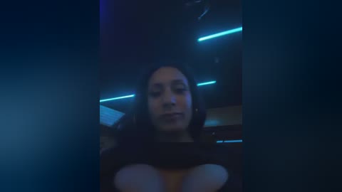 A dimly lit video featuring a woman with long dark hair, wearing a black top, sitting in a dimly lit room with neon blue lights in the background.