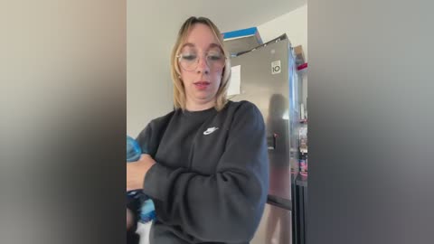 Video of a young Caucasian woman with glasses, fair skin, and shoulder-length blonde hair, wearing a black sweatshirt, holding a blue and white object in a kitchen with stainless steel appliances.