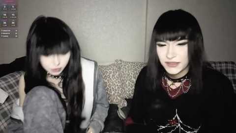Video of two pale-skinned Asian women with long black hair, wearing goth-inspired outfits with chokers and dark makeup, sitting on a patterned couch, with a video game controller in the foreground.