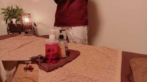 A video of a dimly-lit room with a person in a red and white outfit standing beside a massage table covered with beige sheets. The table is adorned with candles, a bottle of lotion, and a massage tool.