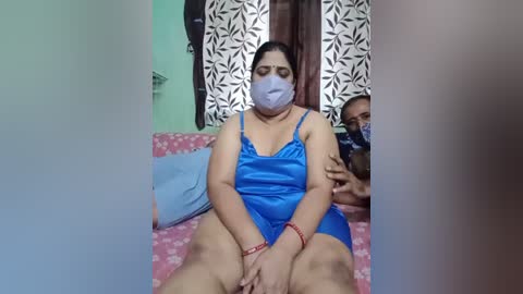 Video of a South Asian woman in a blue satin nightgown, seated on a pink floral bed, wearing a face mask. She has a large build, dark skin, and medium-sized breasts. Her hands cover her crotch.