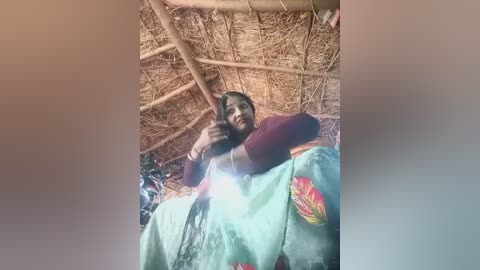 Video of a smiling woman in traditional attire with a red top and floral saree, sitting on a mat inside a thatched hut with straw roof, holding a phone, taking a selfie.