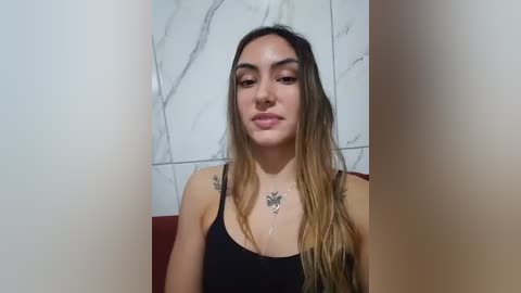 Video of a young woman with long, wavy, light brown hair and fair skin, wearing a black spaghetti-strap top, seated against a white marble tiled wall.