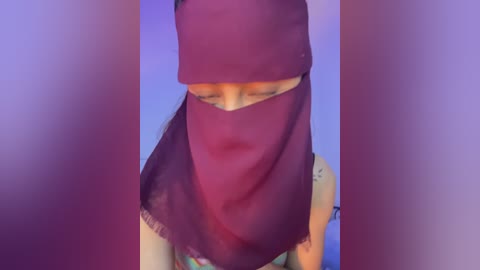A video of a person wearing a maroon hijab with a floral pattern, partially covering their face, against a blurred purple background.