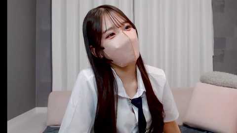 A video of an East Asian woman with long black hair, wearing a white shirt and tie, covering her mouth with a white mask, sitting on a light pink couch in a modern, minimalist room.