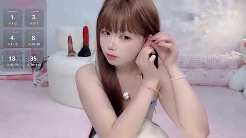 Video of an Asian woman with long brown hair, small breasts, in a pink room, adjusting her hair with a silver bracelet, surrounded by sex toys.