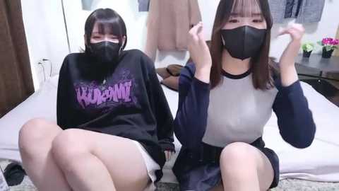 Video of two Asian women in a bedroom; both wear black face masks, black hoodies, and white shorts. One has a pink hoodie with text, the other a navy-blue hoodie. Background includes a bed, white walls, and a brown door.