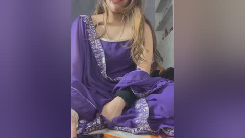 Media: Video of a light-skinned woman with long blonde hair, wearing a purple saree with intricate silver embroidery, seated in a dimly lit room.