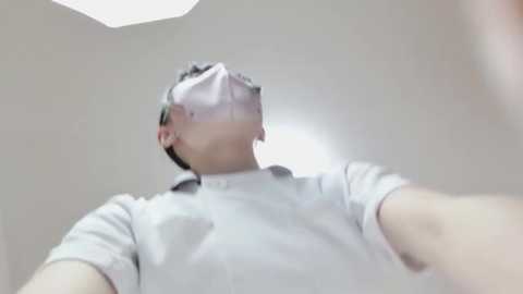 A video of a person wearing a VR headset, standing in a dimly lit room with white walls and a triangular ceiling light. The person is wearing a white t-shirt, and the image is taken from a low angle.