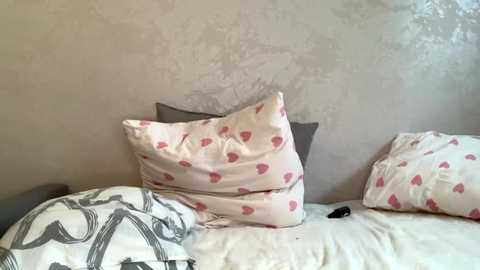 Video of a messy bed with white sheets and pillows featuring pink heart patterns, set against a textured beige wall. The bed has a gray blanket and a black item on the side.