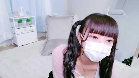 A young Asian woman with long black pigtails and a white surgical mask, wearing a white top, sits on a white bed in a bright, minimalist room with light blue curtains.