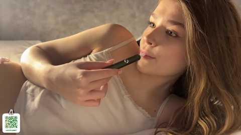 Video of a young, fair-skinned girl with long blonde hair, wearing a white tank top, lying on a bed, holding a black smartphone to her lips, smiling. Background is a blurred, textured wall.