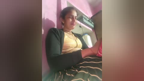 Video of a woman with medium skin tone, dark hair, wearing a black cardigan over a yellow blouse and striped skirt, seated in a pink room with a TV and blue water bottle visible.