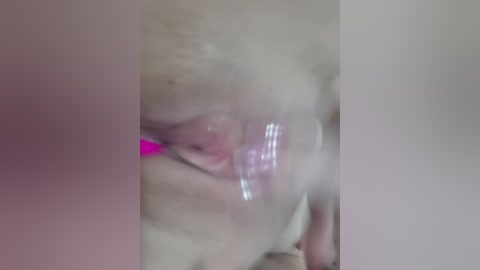 A blurry video of a person's face, possibly female, with visible facial hair, wearing a pink headband, and a pink object, likely a dildo, is inserted into their mouth. The background is out of focus.