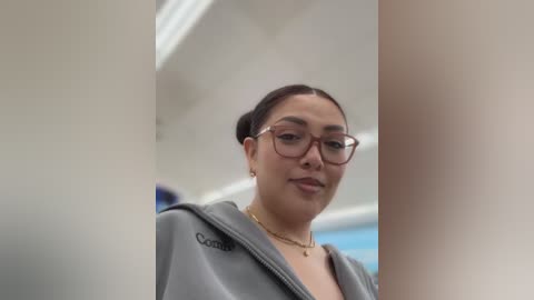 Video of a young woman with medium brown skin, dark hair in a bun, wearing glasses, and a gray Columbia jacket, standing in a modern, white corridor with blurred blue objects in the background.