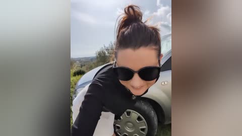 Video of a woman with a high ponytail and sunglasses, leaning into a white car on a sunny day with a blurred background of greenery and a blue sky.