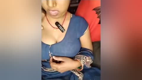 Video of a woman with medium skin tone wearing a blue sari with gold embroidery, holding a black lipstick tube, sitting in a car with red and orange cushions in the background.