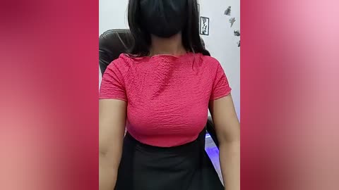 Video of a woman wearing a black mask, red lace top, and black skirt, sitting in a chair against a pink gradient background.