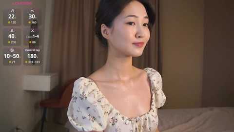 Video of an East Asian woman with fair skin, black hair in a bun, wearing a floral dress, indoors with beige curtains and a bed in the background.