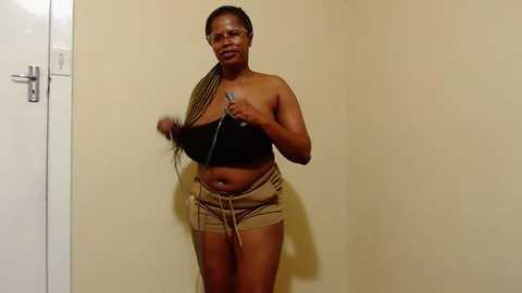Video of a Black woman with a medium-dark complexion, wearing a black bra and beige shorts, adjusting a necklace against a plain beige wall.