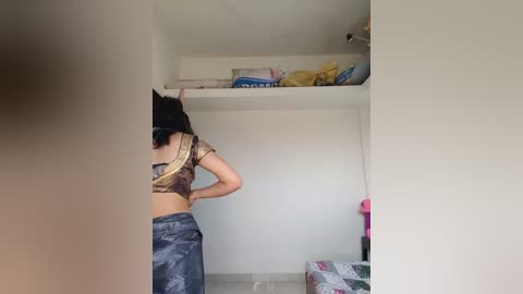 Video of a woman with dark hair wearing a brown crop top and blue sarong, standing in a small, cluttered room with a bed, white walls, and shelves.