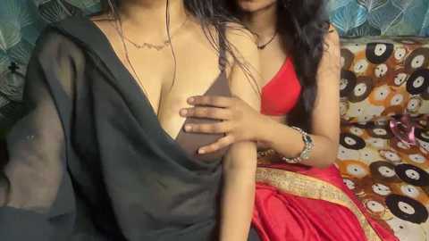 Video of two South Asian women, one partially dressed in a black sari, the other in a red blouse, sitting together on a patterned couch. The woman in the red blouse gently touches the other's breast.