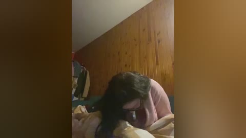 A video of a person with long black hair lying on a bed with beige and brown bedding, wooden wall in the background. The person's head is covered, possibly sleeping.