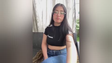 A video of an East Asian woman with long black hair, wearing glasses and a black crop top, seated on a wicker chair with a beige curtain background.