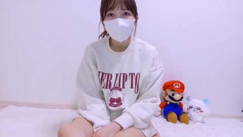 Video of a young Asian woman with short brown hair, wearing a white mask, sweatshirt, and shorts, sitting on a white floor next to Mario and Luigi plush toys.