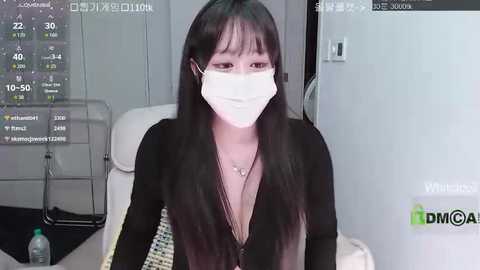 Video of an East Asian woman with long black hair, wearing a black top and white mask, seated in a modern living room.