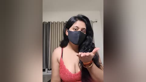 Video of a young woman with long black hair, wearing a red lace bralette, black mask, and blowing a kiss. Background features a window with beige curtains.