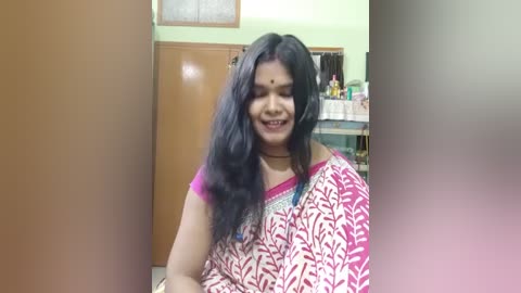Video of a South Asian woman with long black hair, wearing a red and white floral sari with a magenta blouse. She smiles, in a home setting with a wooden door, a framed calendar, and shelves with bottles in the background.