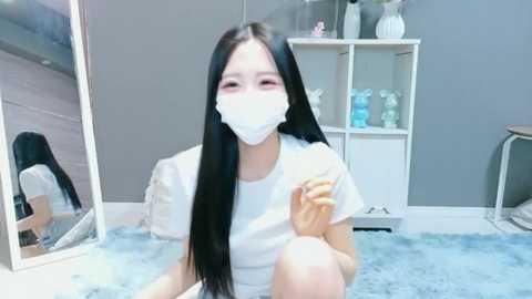 A video of an Asian woman with long black hair, wearing a white mask and T-shirt, sitting on a blue shag rug in a gray room with mirrored walls and shelves filled with white vases.