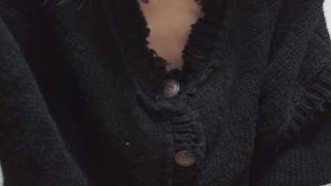 A close-up video of a person's torso wearing a dark, textured cardigan with a frilled neckline and a large, dark button. The background is out of focus.