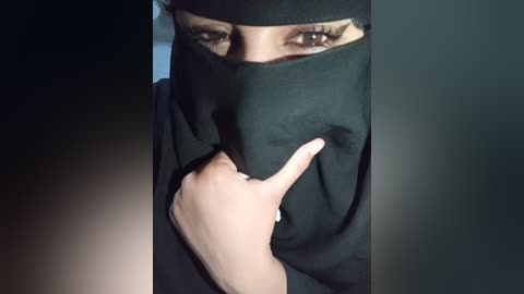 A video of a woman in a black niqab, with her right hand covering her mouth, revealing a hint of a smile. Her brown eyes and dark hair are visible.