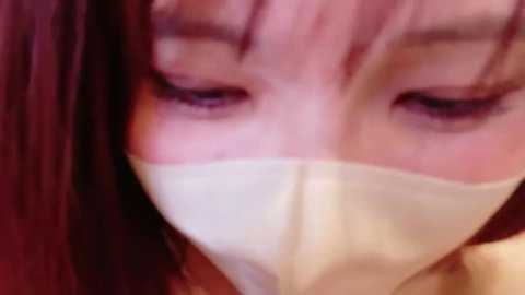 Video of a close-up of a person with light skin, wearing a white mask covering the nose and mouth. The person has long, straight brown hair with bangs. The background is out of focus, with a warm, reddish-brown tone.